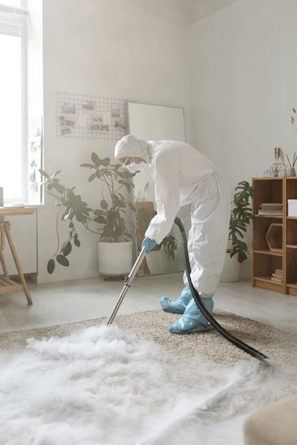 A professional cleaner from Carpet Cleaning Merton is performing deep surface cleaning on a beige carpet in a well-lit, modern living room. The technician is dressed in a full white protective suit, including gloves, a face mask, and shoe covers, using a vacuuming or extraction machine connected to a hose to thoroughly clean the carpet. The room features white walls, a large window allowing natural light, and indoor plants in white ceramic pots, with a shelving unit containing decorative items and a white table beside the window. The clean, tidy space highlights the effectiveness of professional sanitisation and deep cleaning processes for maintaining hygienic and fresh household surfaces.
