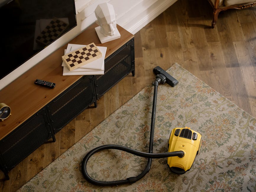 A yellow vacuum cleaner with a black hose and silver extension wand is positioned on a patterned area rug in a living room. To the left, a wooden TV stand with black metal legs holds a remote control, a stack of papers, and a white bust sculpture. The room features hardwood flooring and white skirting boards, with natural light illuminating the space, highlighting the cleanliness and well-maintained surfaces, emphasizing the importance of surface cleaning and deep cleaning services provided by Carpet Cleaning Merton near Wimbledon Common.