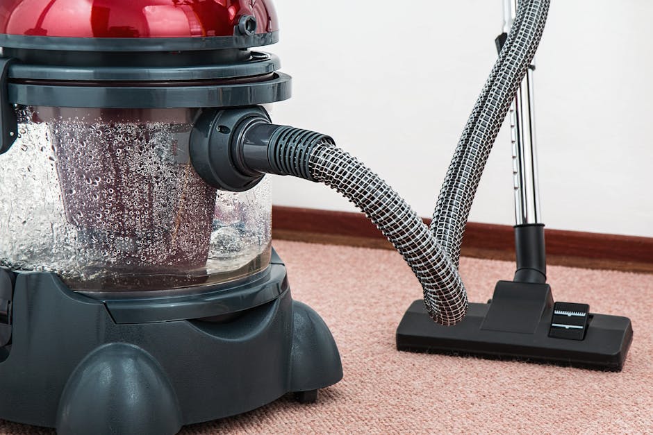 A professional cleaner from Carpet Cleaning Merton is performing deep surface cleaning on a beige carpet in a well-lit, modern living room. The technician is dressed in a full white protective suit, including gloves, a face mask, and shoe covers, using a vacuuming or extraction machine connected to a hose to thoroughly clean the carpet. The room features white walls, a large window allowing natural light, and indoor plants in white ceramic pots, with a shelving unit containing decorative items and a white table beside the window. The clean, tidy space highlights the effectiveness of professional sanitisation and deep cleaning processes for maintaining hygienic and fresh household surfaces.