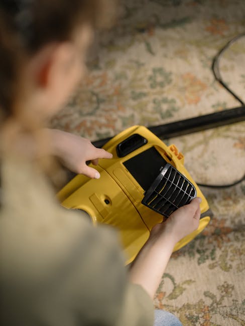 Close-up view of a person operating a yellow portable carpet cleaning or sanitisation device, focusing on their hands and the equipment. The device is held over a traditional patterned area rug with muted tones of beige, orange, and green, situated on a wooden floor. The background features part of the patterned rug and a black power cord connected to the equipment. The scene is well-lit, highlighting the cleanliness of the rug and the equipment used for surface cleaning or deep cleaning, aligning with services offered by Carpet Cleaning Merton for domestic cleaning on Mitcham High Street, Merton.