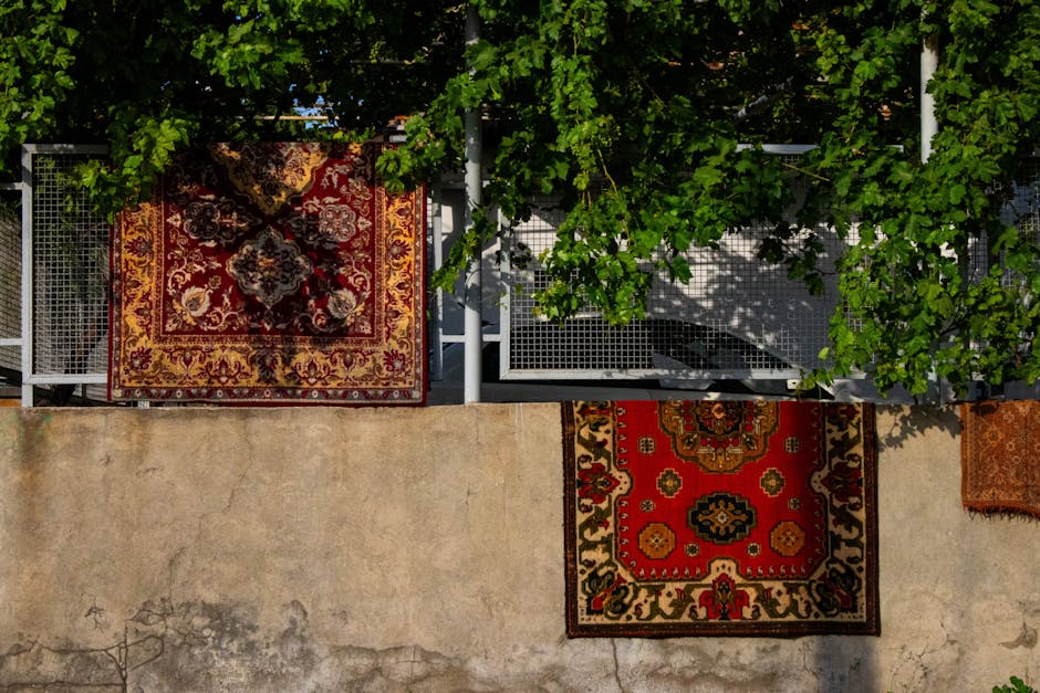 An aerial view of an outdoor balcony area featuring two Persian-style rugs with intricate patterns in red, cream, and black hues. The balcony has a metal railing and is shaded by lush green leaves from a nearby tree, creating dappled sunlight on the surface. The flooring is a light, weathered concrete with some stains visible. In the background, there is a section of a rooftop terrace with a white metal grid and a partially visible outdoor chair. The overall scene emphasizes surface cleaning and maintenance in a residential setting, fitting with the services offered by Carpet Cleaning Merton for deep cleaning and sanitisation.