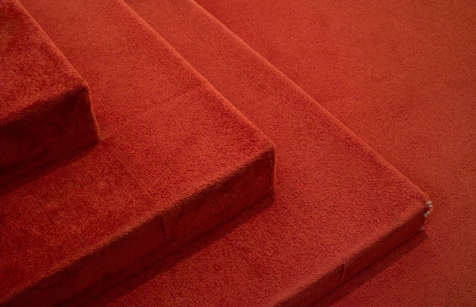 Close-up image of a section of a red carpeted staircase in a residential or commercial setting. The staircase features soft-textured, deep red carpet with visible fibers, covering several steps with a uniform appearance. The top step shows a slight damage at the edge, revealing a small tear or fray in the carpet fibers. The surrounding area is well-lit, highlighting the plush surface and consistent color. The carpet appears clean and well-maintained, with no visible stains or dust, indicative of thorough surface cleaning or maintenance by Carpet Cleaning Merton, a professional cleaning service specializing in domestic and commercial hygiene solutions.