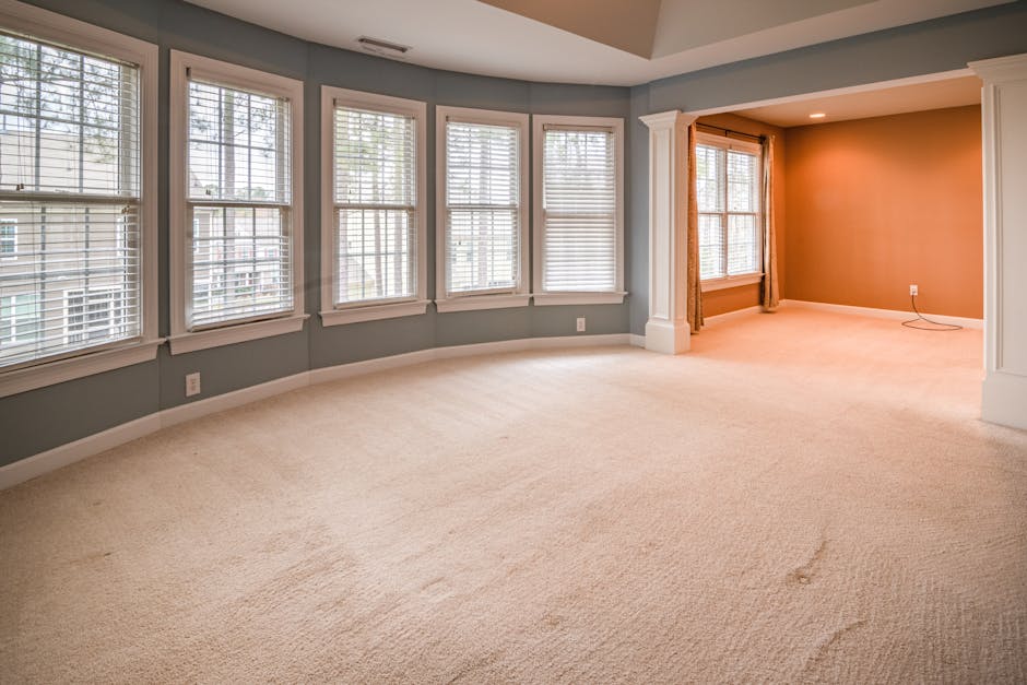 A spacious living room with light beige carpet flooring that appears clean and well-maintained. The room features multiple large white-framed windows with white blinds, allowing natural light to fill the space. The walls are painted in soft shades of blue and beige, creating a bright and welcoming atmosphere. The room includes decorative white columns and trim around the windows and an open archway leading to another area, which showcases polished wooden window frames and a subtle orange accent wall. The overall appearance is tidy, with no visible dust or clutter, reflecting professional surface cleaning and upkeep by Carpet Cleaning Merton as part of their domestic cleaning services.