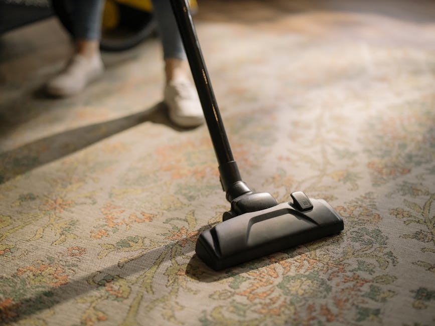 Close-up of a black vacuum cleaner head actively cleaning an ornate, patterned carpet in a home setting, with a person in casual shoes and jeans visible in the background. The carpet features intricate floral designs in pastel colors on a beige base, appearing clean and well-maintained. The vacuum is positioned in the center of the image, with a slight tilt, highlighting its motorized brush head. Soft natural lighting illuminates the area, emphasizing the cleanliness of the surface. The setting suggests professional surface cleaning and deep cleaning services provided by Carpet Cleaning Merton, aligned with domestic cleaning and sanitisation activities in Wimbledon Park flats.