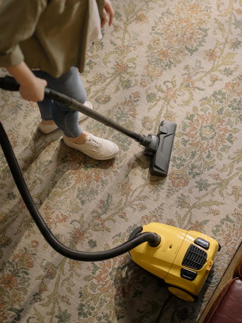 A person wearing beige sneakers, blue jeans, and a beige jacket is operating a vacuum cleaner over a patterned area rug in a room with soft, natural lighting. The vacuum cleaner is yellow with a black hose and attachment, and is positioned on a floral-designed rug with beige, green, and orange tones. The person is holding the vacuum's handle, guiding it across the surface, indicating a deep cleaning or surface cleaning process. The room appears tidy, with a focus on maintaining cleanliness of the rug, aligning with domestic cleaning services provided by Carpet Cleaning Merton for Wimbledon Park flats and other residential spaces.